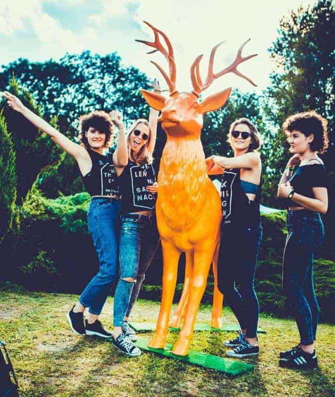 Cerf Jägermeister 2 Cztery kobiety pozują przy pomarańczowej figurze jelenia na tle zieleni.