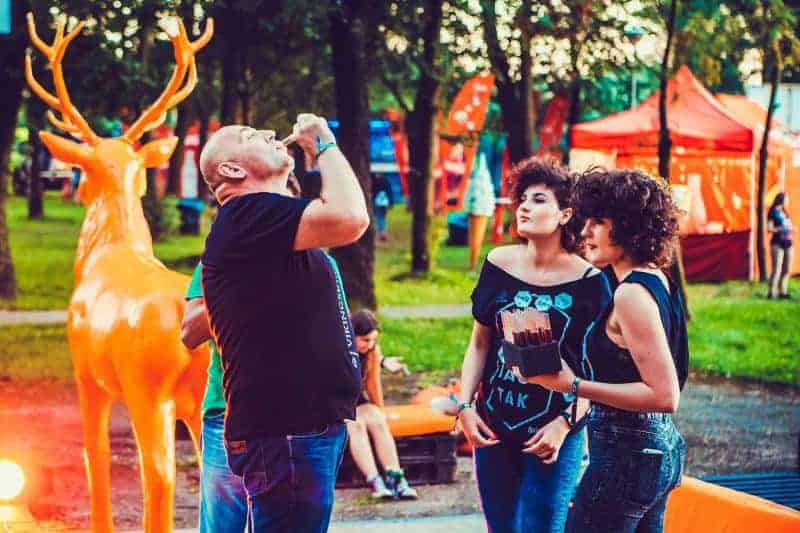 Cerf Jägermeister 4 Ludzie na festiwalu na świeżym powietrzu, z dużą pomarańczową figurą jelenia w tle, w otoczeniu drzew i stoisk.