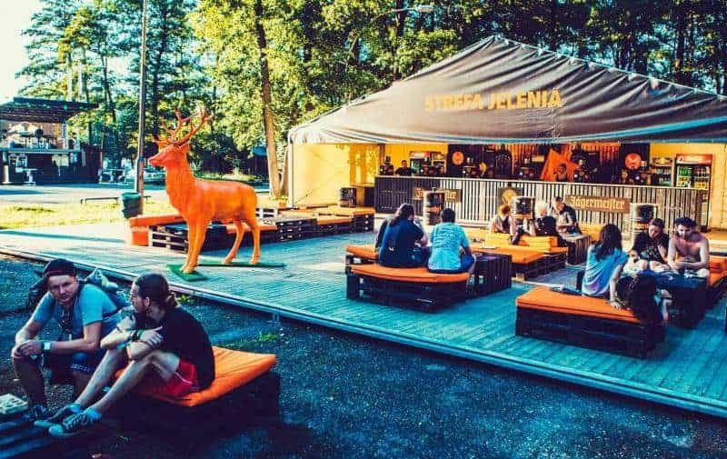 Cerf Jägermeister 1 Ludzie siedzący na pomarańczowych poduszkach na drewnianej platformie przed namiotem z napisem