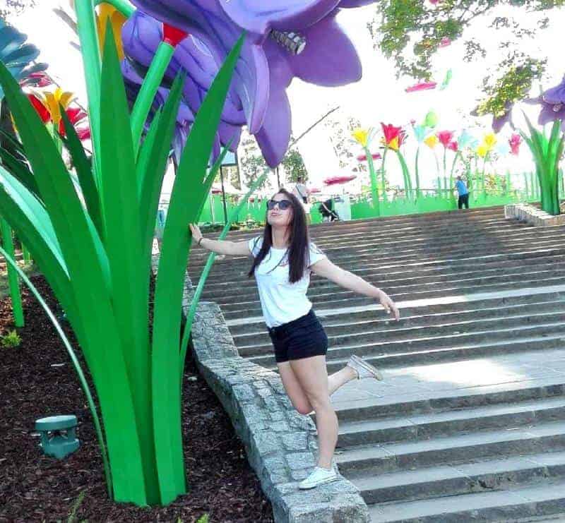 Fleur de Legendia 4 Kobieta pozująca przy gigantycznych modelach kwiatów w parku.