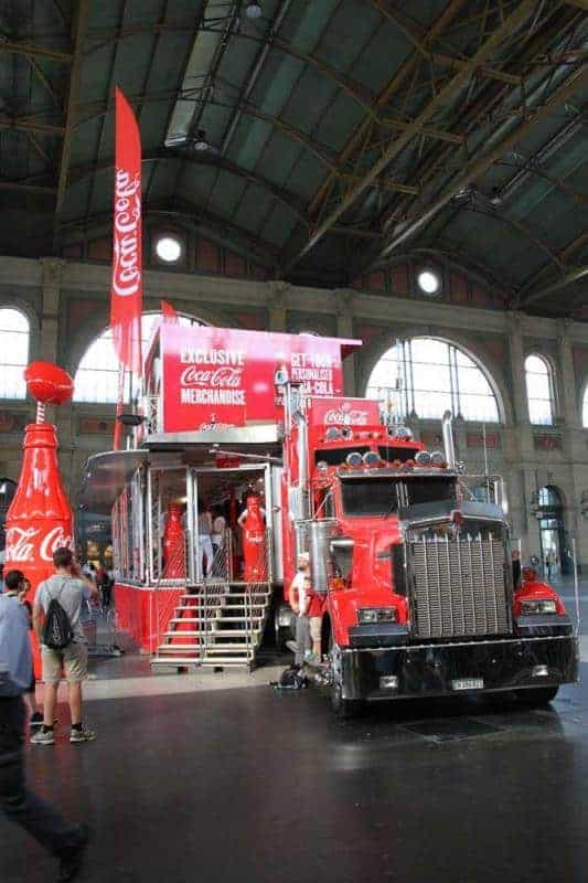 100e anniversaire de la bouteille en verre Coca-Cola 3 Czerwony ciężarówka Coca-Cola z naczepą przekształconą w stoisko sprzedażowe z produktami Coca-Cola, stojąca w dużej hali.