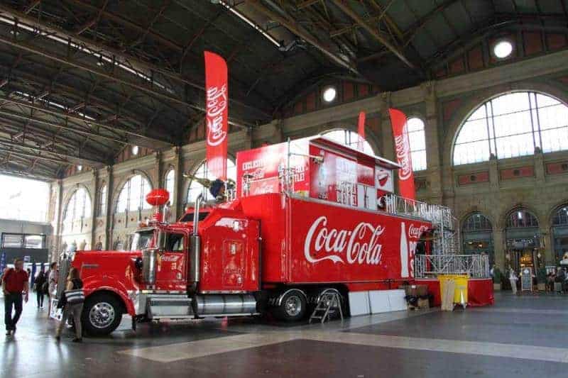 100e anniversaire de la bouteille en verre Coca-Cola 4 Czerwony ciężarówka Coca-Cola w hali dworca z dużymi flagami Coca-Cola i dużymi oknami w tle.