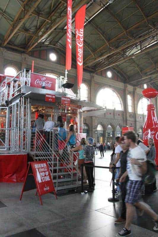 100e anniversaire de la bouteille en verre Coca-Cola 2 Stoisko Coca-Coli z ludźmi w hali dworca kolejowego.