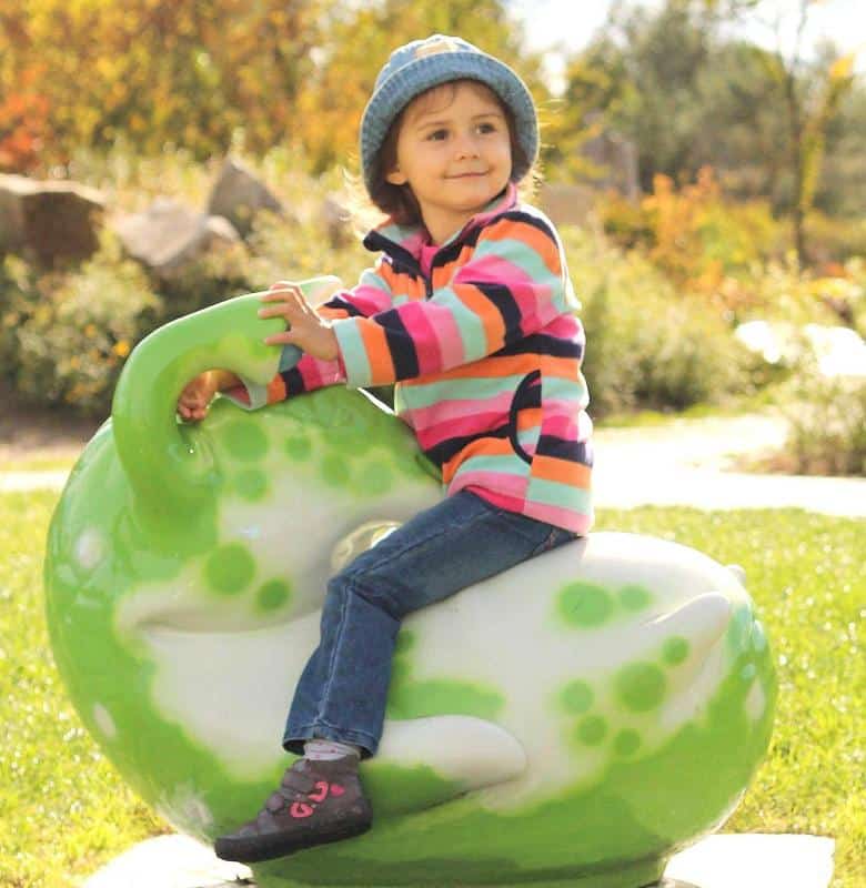 Figurines dans le parc d'attractions 4 Dziecko w kolorowej bluzie i niebieskiej czapce siedzi na zielonej huśtawce w kształcie dinozaura na placu zabaw.