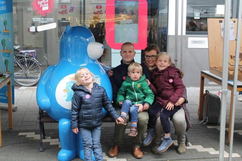 Ours sur un Banc 2 Rodzina z trójką dzieci pozuje do zdjęcia obok dużej niebieskiej maskotki w przestrzeni publicznej.