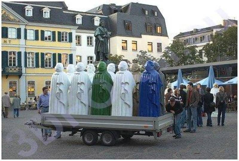 Réplique du monument Beethoven 2 Grupa kolorowych rzeźb na przyczepie na placu miejskim, budynki w tle.