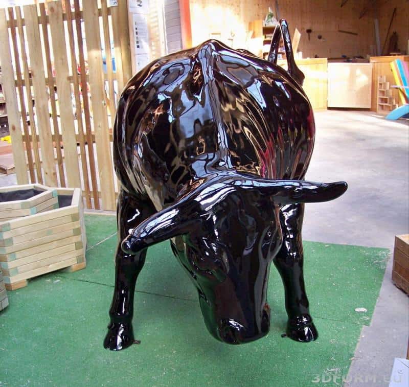 Czarna, błyszcząca rzeźba byka w pozycji pochylonej do przodu, umieszczona na zielonej sztucznej trawie w pomieszczeniu.