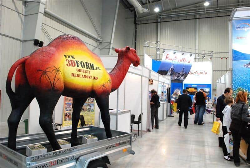 Maquette mobile de chameau 4 Stoisko wystawowe z dużą figurą wielbłąda promującą obiekty reklamowe 3D; w tle kilka stoisk z banerami i ludzie oglądający ekspozycje.