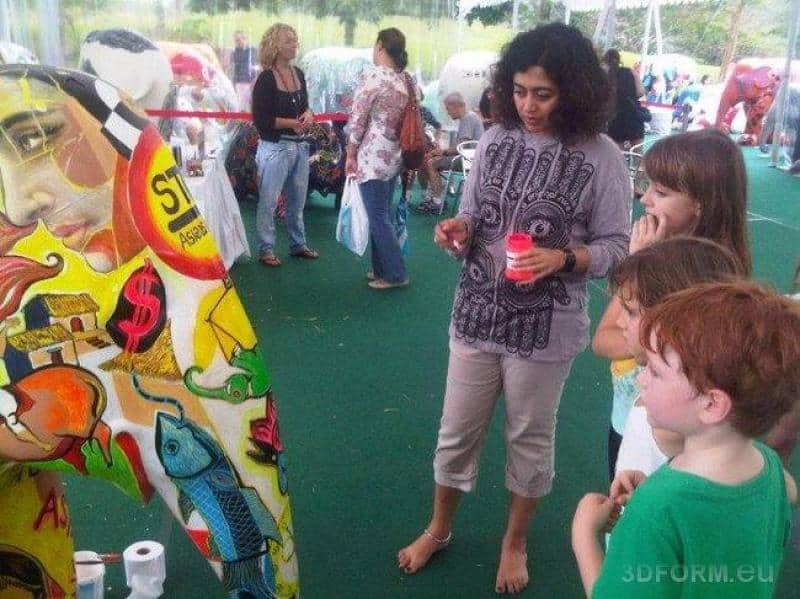 Grandes figures peintes par des enfants 2 Kobieta i dzieci stojące przed kolorowym muralem na wystawie plenerowej.