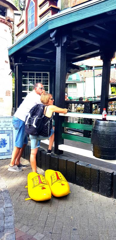 Grands Sabots 3 Para osób w żółtych, dużych drewnianych butach pozuje do zdjęcia przed budynkiem w stylu holenderskim.