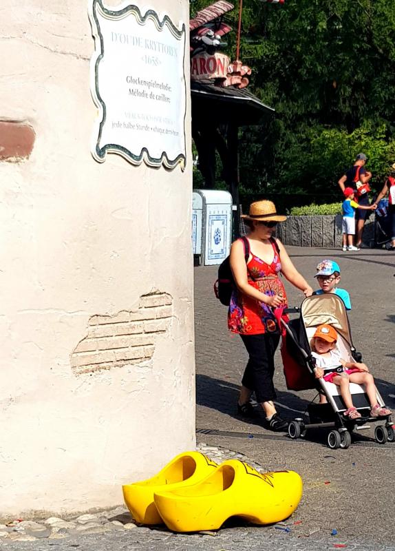 Grands Sabots 4 Kobieta z wózkiem dziecięcym przechodzi obok dużych żółtych drewniaków przy ścianie z francuskim napisem.