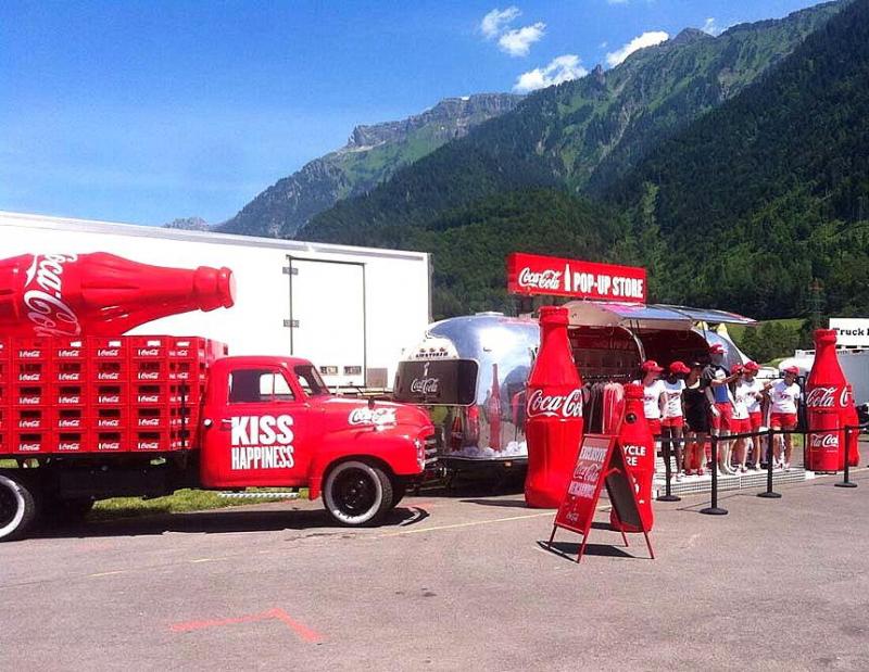 Camion publicitaire Coca-Cola 3 Czerwony samochód Coca-Coli z dużą butelką na przyczepie i sklepem pop-up w tle, stojący na parkingu w otoczeniu gór.