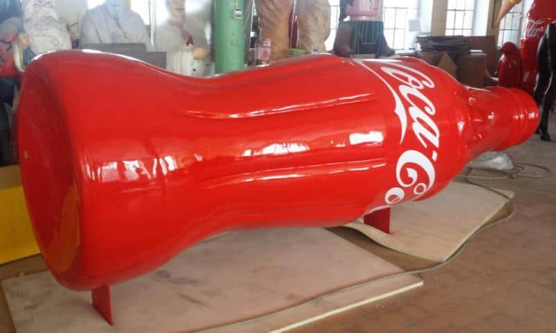 Camion publicitaire Coca-Cola 2 Duża rzeźba w kształcie butelki Coca-Coli w czerwonym kolorze, znajdująca się w warsztacie.