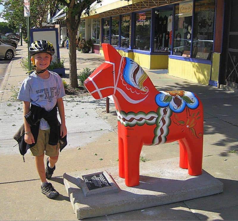 Chevaux de Dalécarlie artistiquement peints 2 Chłopiec w kasku na rower stojący obok kolorowej rzeźby konia na chodniku wzdłuż ulicy z witrynami sklepów.