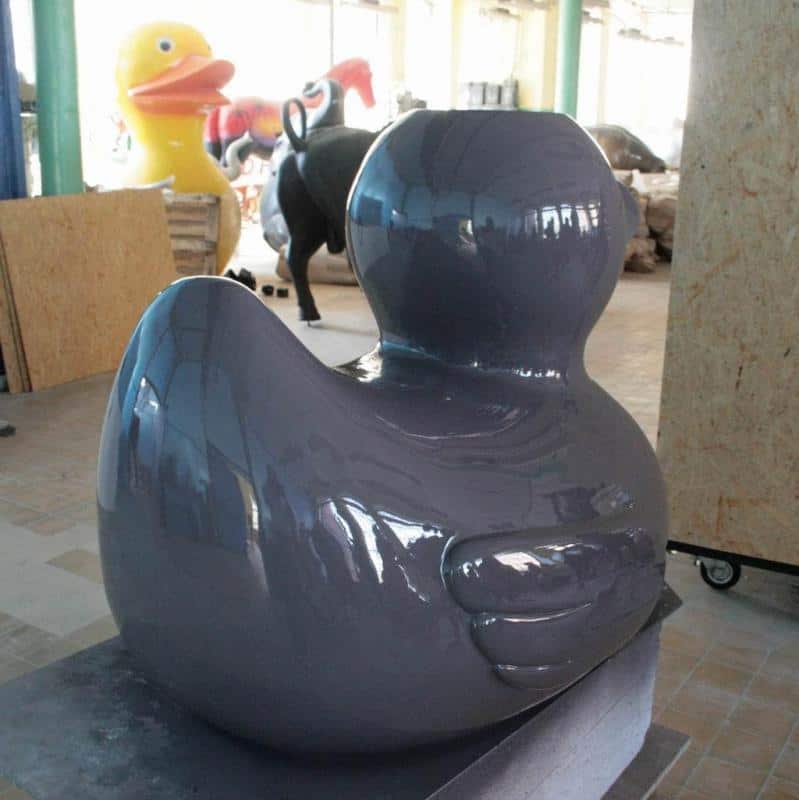 Attractions du festival 3 Duża, szara rzeźba kaczki w warsztacie z większymi rzeźbami w tle, w tym żółtą kaczką.