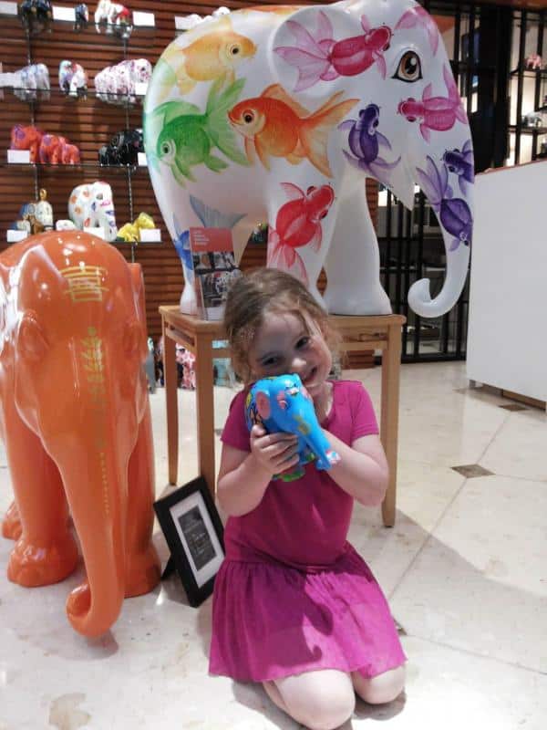 Dziewczynka w różowej sukience, trzymająca kolorową figurkę słonia, siedząca przy większych, pomalowanych słoniach dekoracyjnych.