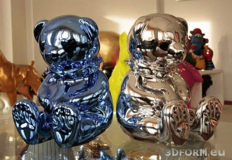 Figures métallisées, chromées 3 Dwie błyszczące figurki misiów, jedna w kolorze niebieskim, druga srebrna, stojące na szklanej powierzchni.