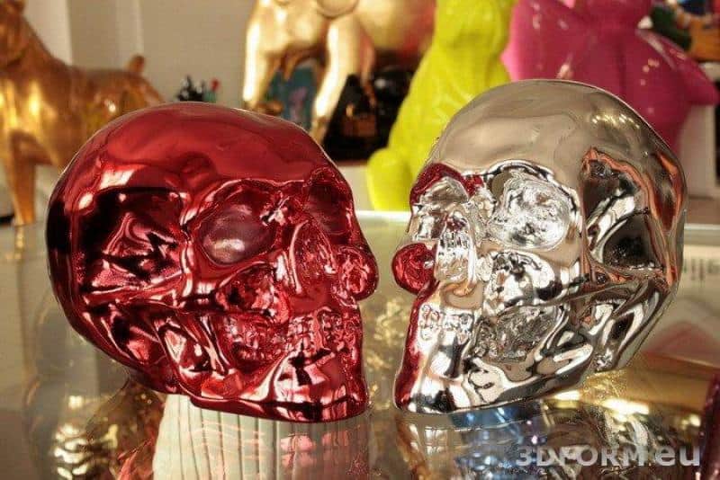 Figures métallisées, chromées 4 Dwie metaliczne czaszki, jedna czerwona, druga srebrna, ustawione na szklanym stole.