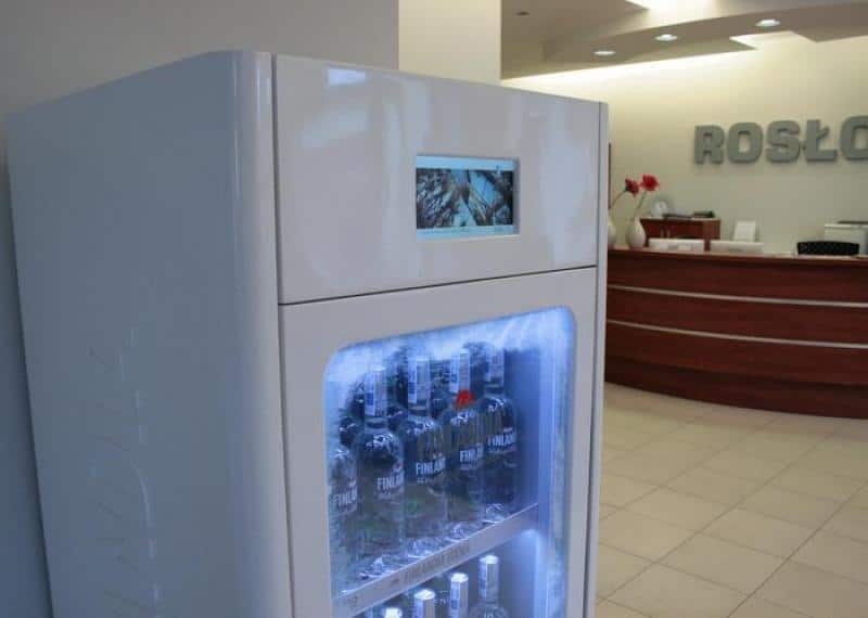 Lodówka z napojami w recepcji firmy. W tle biurko recepcyjne i neonowy napis na ścianie.