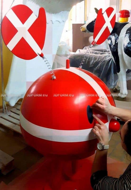 Figurine publicitaire Hoptimist 1 Czerwona, okrągła figura z uszami na sprężynach i białymi paskami, z rękami przymocowującymi oko. W tle plastikowa krowa.