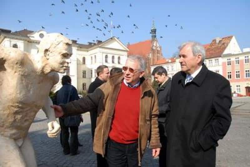 Promotion des Championnats du Monde IAAF 1 Dwóch mężczyzn rozmawiających obok rzeźby na rynku miejskim, w tle kamienice i wieża zegarowa.