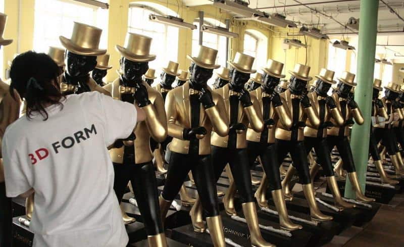 Grandes Figures Johnnie Walker 4 Rzędy manekinów ubrane we złote garnitury i cylindry, osoba w białej koszulce z napisem