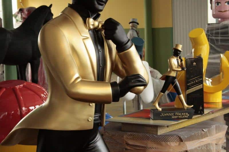 Grandes Figures Johnnie Walker 1 Złota figurka Johni Walker w złotym garniturze, obok mniejsza statuetka i pudełko.