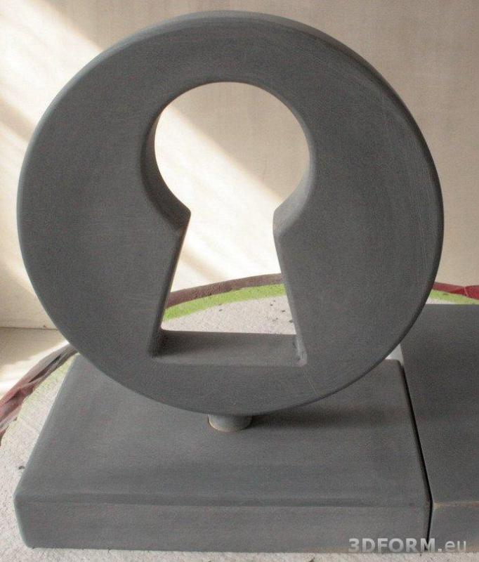 Serrure / Statuette 2 Szara rzeźba w kształcie zamka do klucza, umieszczona na prostokątnej podstawie.