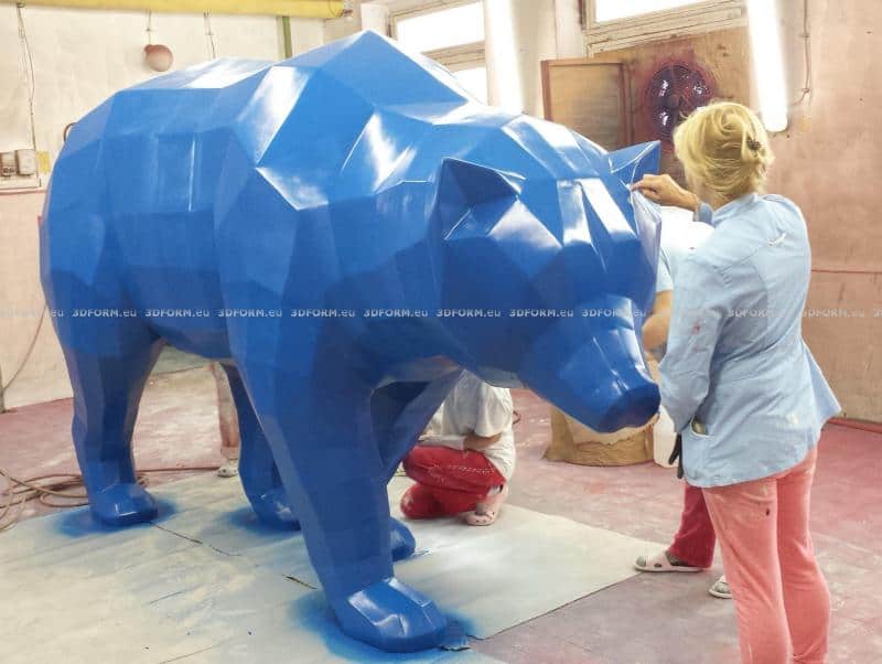 Ours géométrique low-poly 1 Kobieta maluje dużą, niebieską, geometryczną rzeźbę niedźwiedzia w pracowni artystycznej.