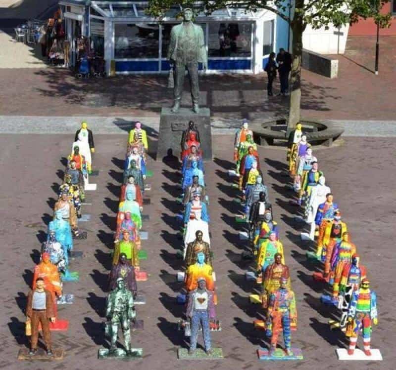 Sculptures de Mineurs 1 Kolorowe figury ludzi ustawione w szeregu na placu przed dużym pomnikiem mężczyzny.