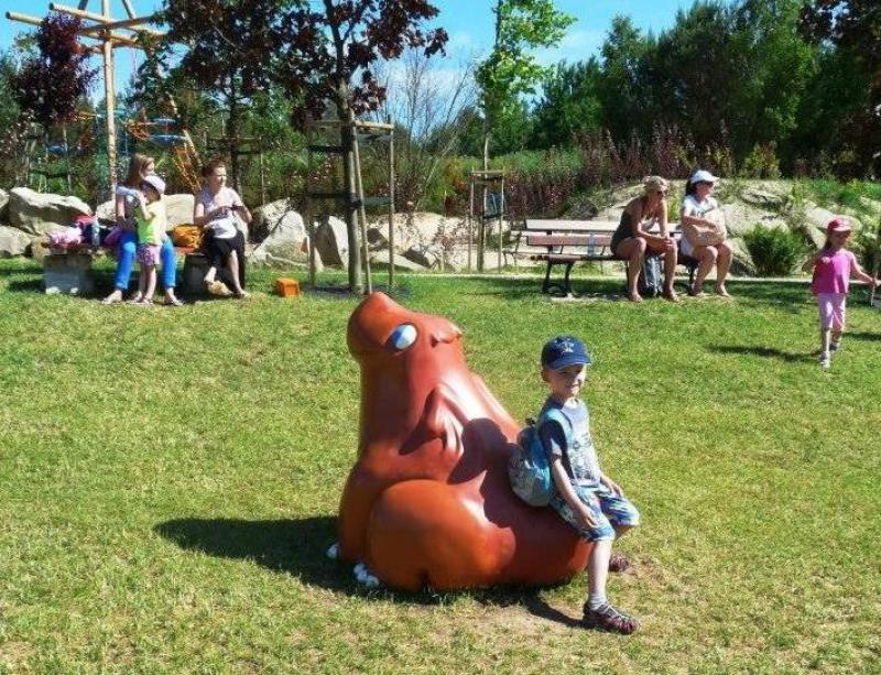Chłopiec siedzący na rzeźbie zwierzęcia w parku, w tle kilka osób i drzewa.