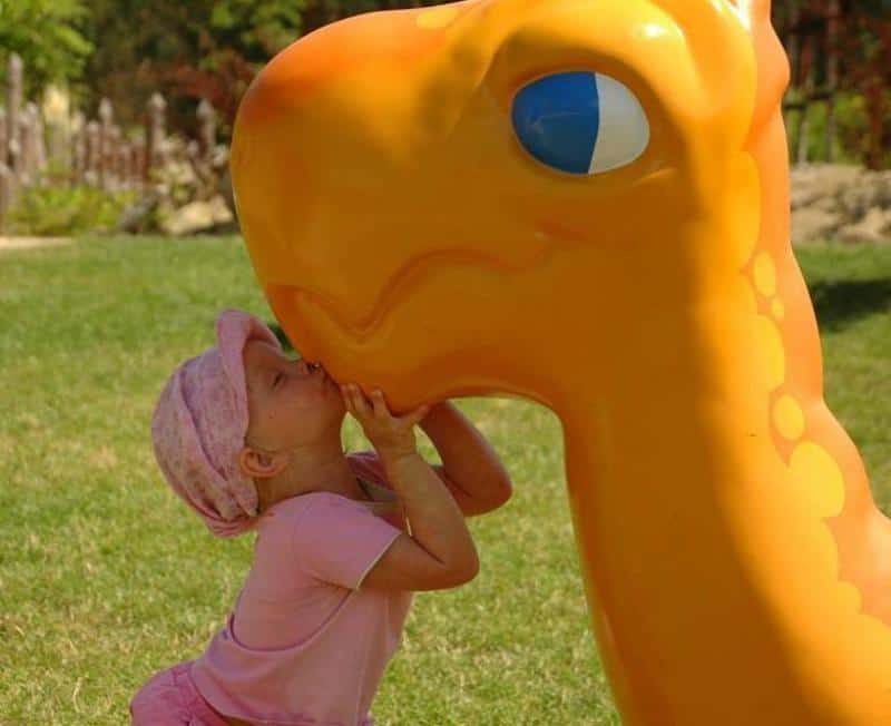 Dziewczynka w różowym ubraniu całuje dużą, pomarańczową figurkę dinozaura na placu zabaw.