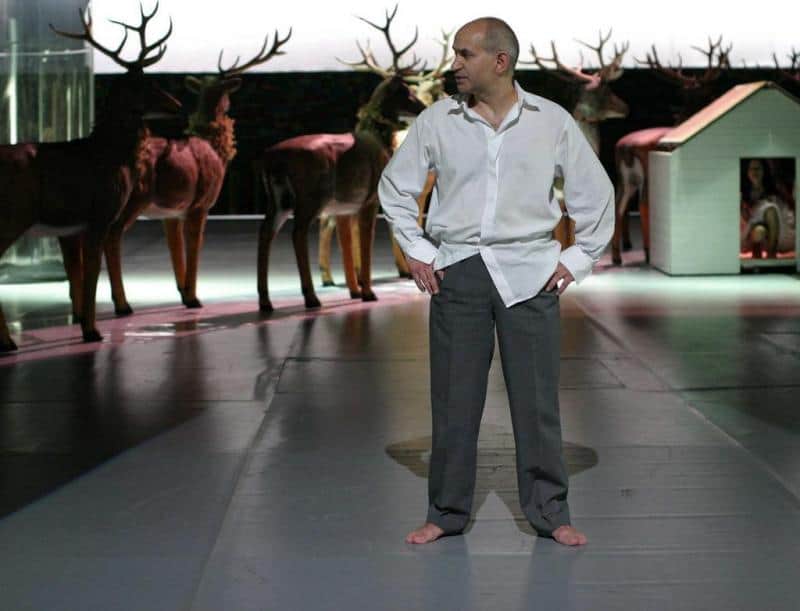 Éléments de scénographie pour le Théâtre Dramatique de la Ville Capitale de Varsovie 1 Mężczyzna w białej koszuli i szarych spodniach stoi boso na scenie, w tle stoją wypchane jelenie.