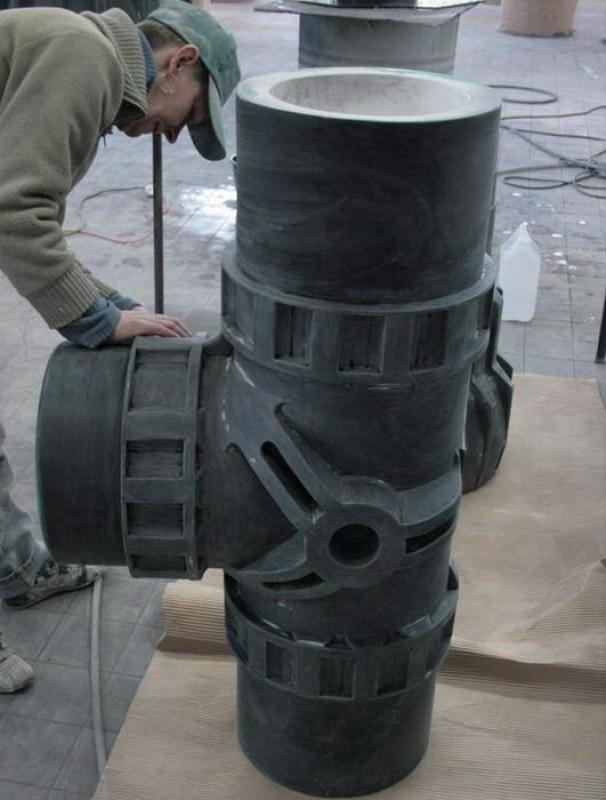 Té - Raccord de Tuyauterie 3 Mężczyzna w warsztacie pochylający się nad dużym rzeźbionym modelem krzyżowego połączenia rur.