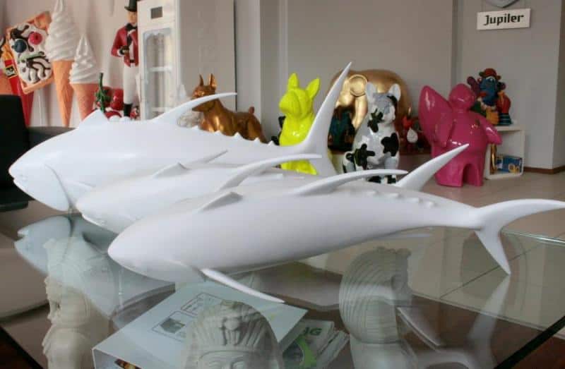 Banc de thons 3 Białe figurki rekinów na szklanym stole, w tle kolorowe figurki zwierząt i inne dekoracje.