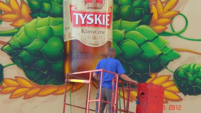 Mężczyzna na podnośniku malujący mural z butelką piwa Tyskie Klasyczne i chmielami.