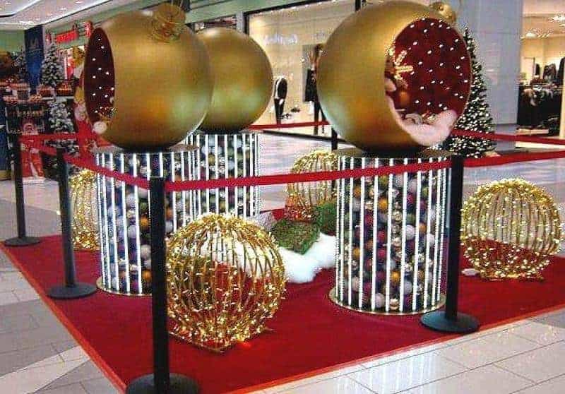Boules de Noël éclairées 4 Duże złote bombki świąteczne stojące na cylindrycznych podstawach, otoczone czerwoną taśmą w centrum handlowym.