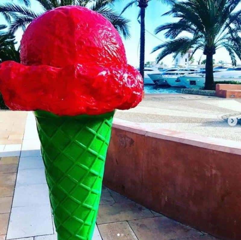 Maquette de glace - La Cabane Verte 4 Wielka plastikowa rzeźba przedstawiająca loda rożka z czerwoną gałką na zielonym wafelku, umieszczona na zewnątrz w otoczeniu palm i jachtów.