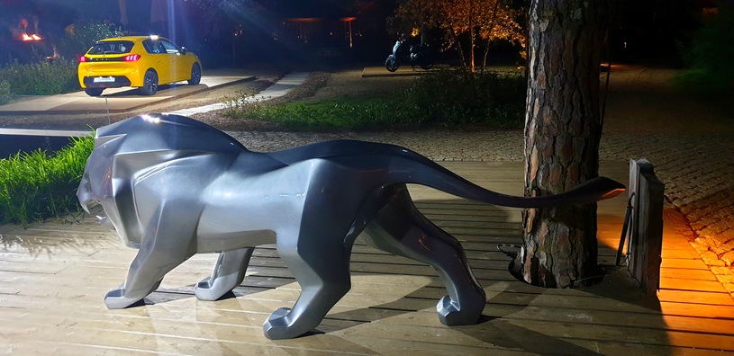 Lion Peugeot 4 Figurka lwa w kolorze srebrnym na drewnianej platformie, w tle żółty samochód na oświetlonym podjeździe.