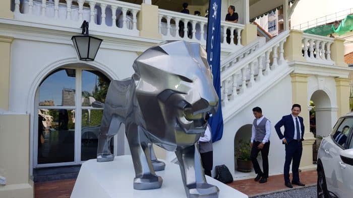 Lion Peugeot 3 Metalowy posąg lwa na tle białego budynku z kolumnadą i schodami, przy którym stoją elegancko ubrani ludzie.