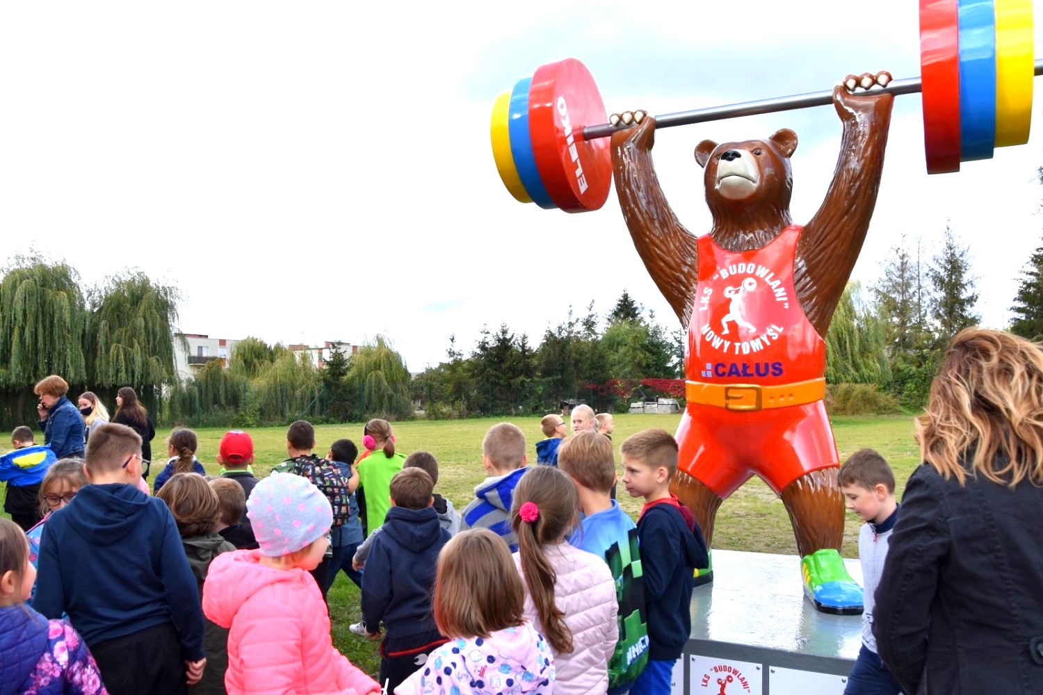 Dzieci z nauczycielkami oglądają dużą figurę misia podnoszącego sztangę na zewnątrz.