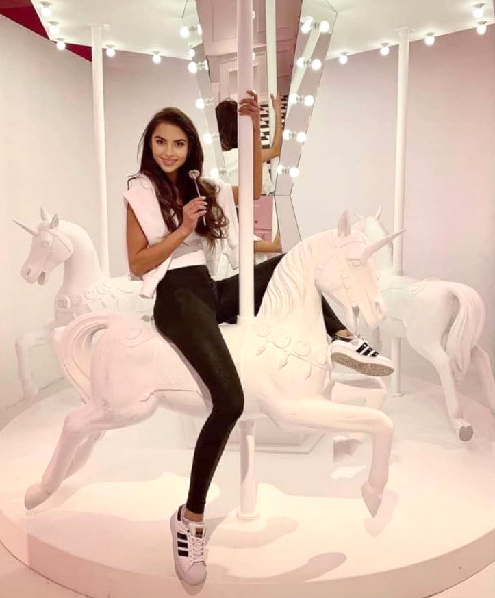 Kobieta siedząca na białym jednorożcu na karuzeli, trzymająca lizaka, otoczona jasnymi światłami.