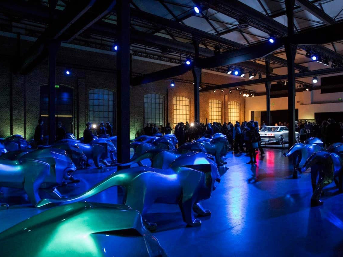 Lion Peugeot 2 Pomieszczenie wypełnione niebieskimi rzeźbami lwów, z ludźmi i samochodem w tle, oświetlone niebieskim i fioletowym światłem.