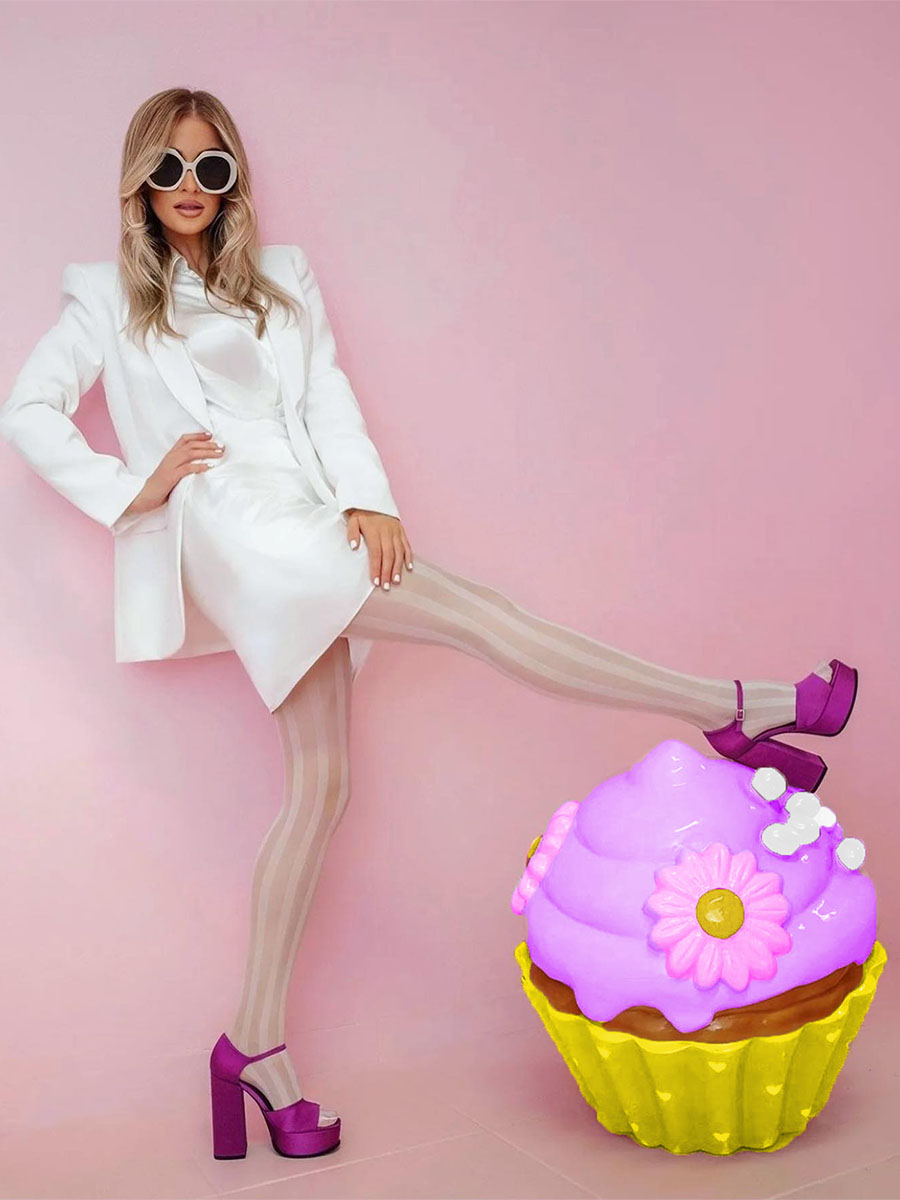 Kobieta w białym stroju, okularach przeciwsłonecznych i fioletowych butach na wysokim obcasie na tle różowej ściany, z fioletowo-żółtą dekoracyjną babeczką.