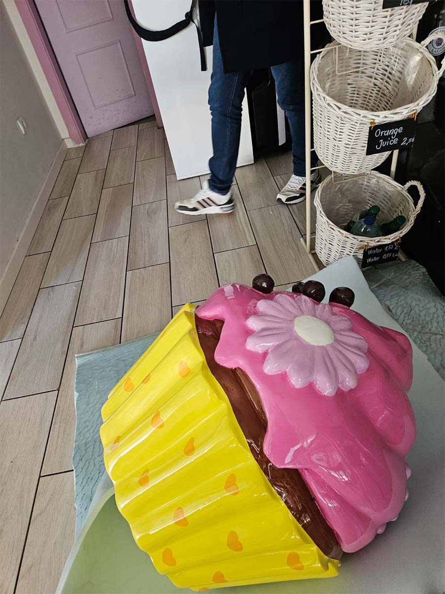 Rzeźba wielkiego babeczki w pastelowych kolorach stojąca na podłodze w kawiarni obok osoby w dżinsach i białych butach.