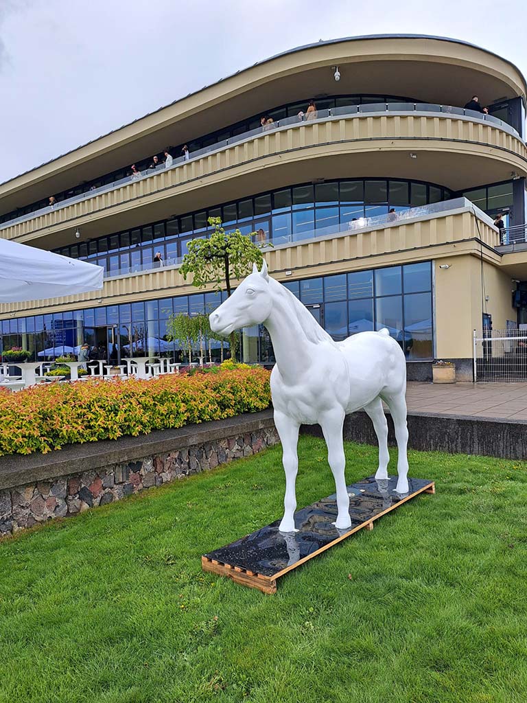 Biały pomnik konia na trawniku przed nowoczesnym budynkiem z dużymi oknami.