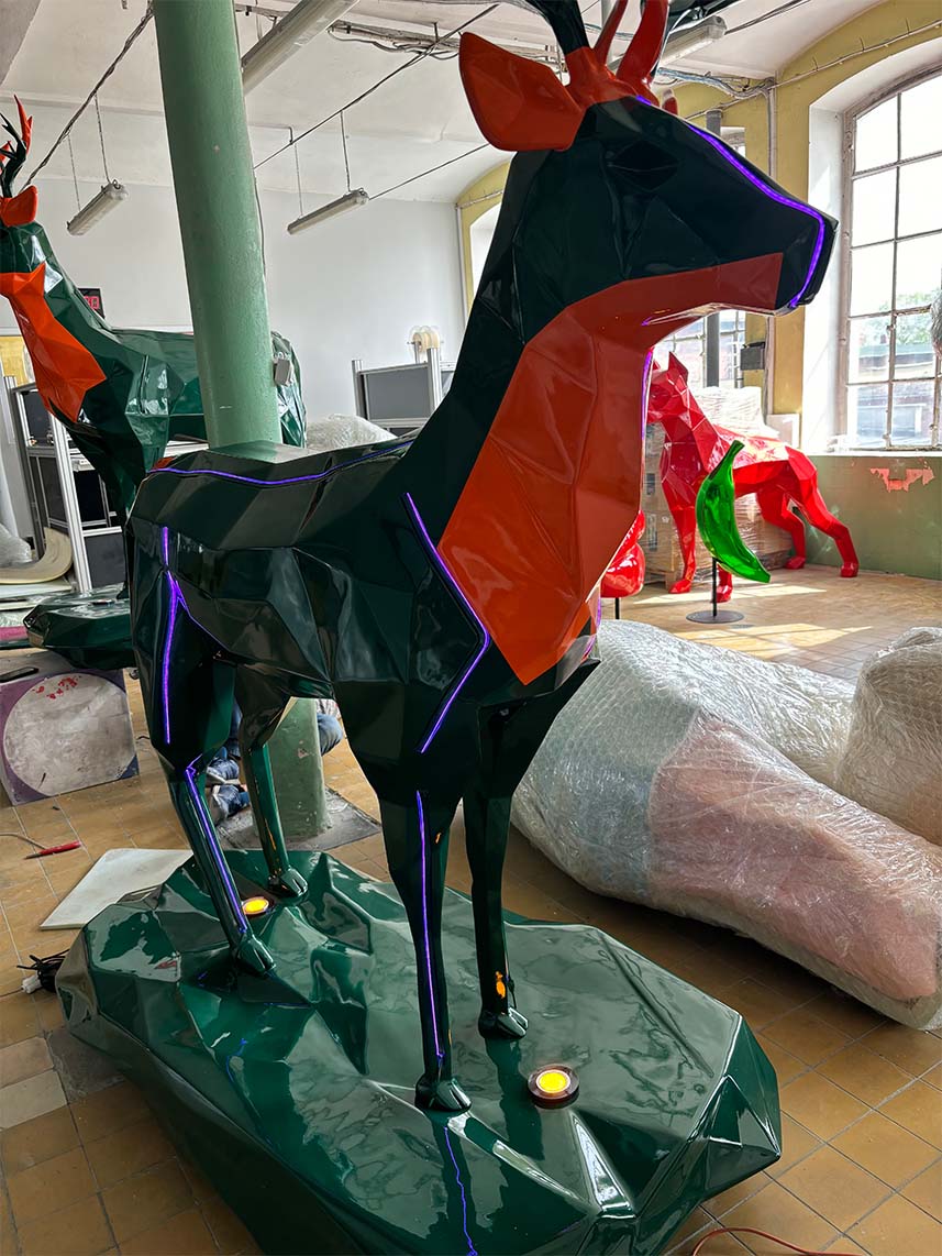 Duży, kolorowy posąg jelenia wewnątrz budynku, otoczony innymi podobnymi rzeźbami i pakunkami.