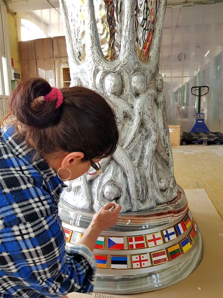 Kobieta w niebiesko-czarnej koszuli w kratę pracuje nad dużą srebrną rzeźbą z motywem ludzkich sylwetek, umieszczoną na podstawie ozdobionej flagami różnych państw.