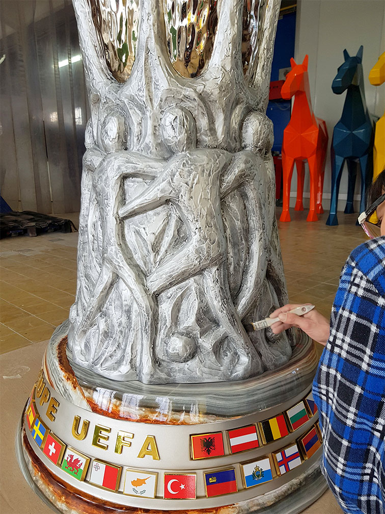 Postać malująca rzeźbę przedstawiającą ludzi grających w piłkę nożną na podstawie z flagami krajów i napisem EUROPE UEFA, we wnętrzu z kolorowymi rzeźbami koni w tle