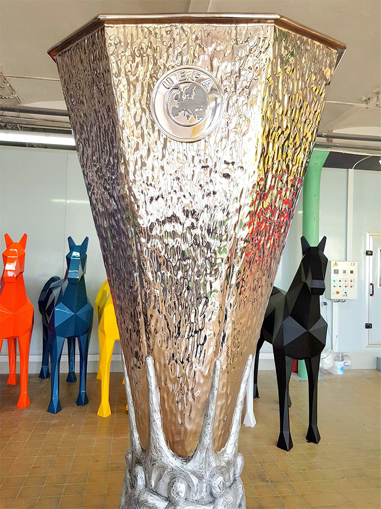 Duży srebrny puchar UEFA stojący na podłodze w pomieszczeniu z mozaikową podłogą, w tle kolorowe geometryczne rzeźby koni w kolorach pomarańczowym, niebieskim, żółtym i czarnym.